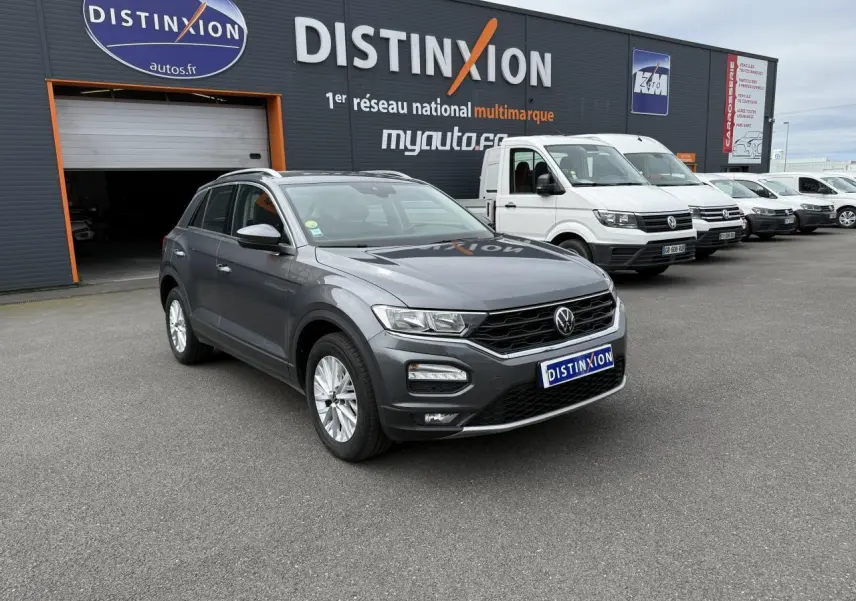 Volkswagen T-Roc gris Indium Metal en 3/4 avant droit devant un bâtiment Distinxion avec plusieurs véhicules utilitaires blancs.
