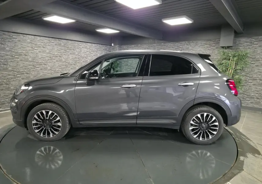 Vue de profil côté gauche d'un Fiat 500X gris avec jantes alliage 17 pouces dans un showroom intérieur.