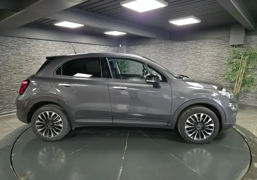 Profil droit d'un Fiat 500X gris 2024 avec jantes alliage 17'' et rétroviseurs noirs dans un showroom.
