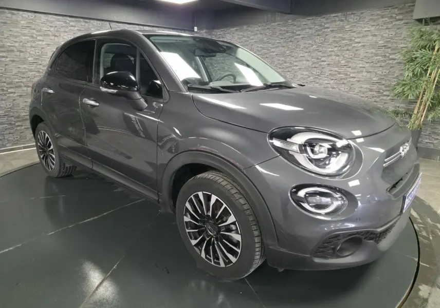 Fiat 500X gris en 3/4 avant droit, avec jantes alliage 17'' et feux LED distinctifs dans un showroom.