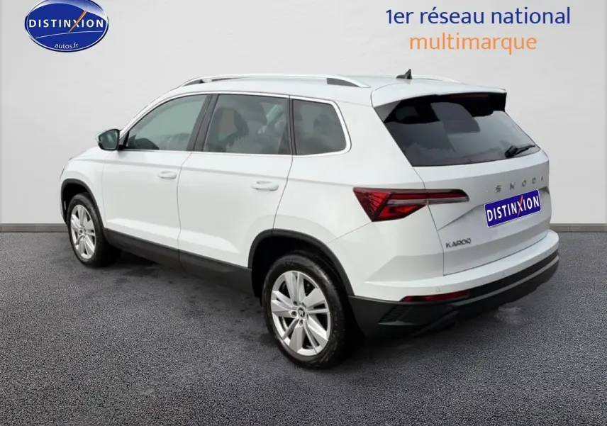Vue 3/4 arrière droite d'un Skoda Karoq blanc glacial avec jantes alu et barres de toit visibles.