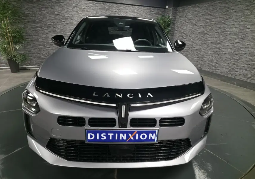 Vue avant d'une Lancia Ypsilon 2025 gris métallisé avec calandre noire et toit bi-color noir en intérieur showroom.