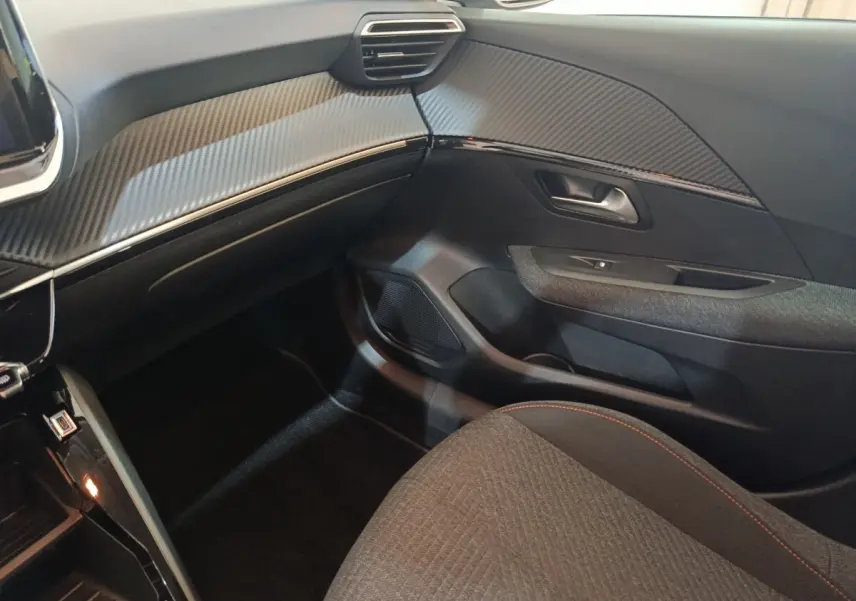 Vue intérieure côté passager de la Peugeot 208 Hybrid 2025, tableau de bord et porte avec finition carbone gris clair.