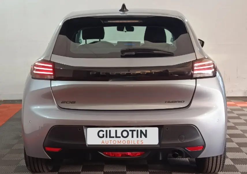 Vue arrière d'une Peugeot 208 Hybrid gris clair avec feux LED allumés sur fond intérieur neutre.