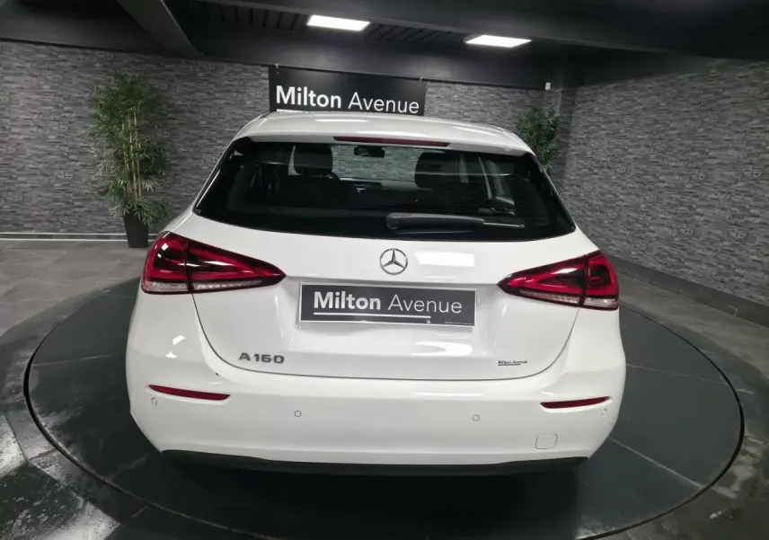 Vue arrière d'une Mercedes Classe A 160 blanche avec feux LED et logo Mercedes visible sur fond intérieur showroom.
