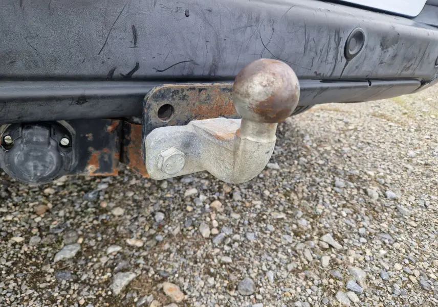 Gros plan sur l'attelage rouillé fixé au pare-chocs arrière noir du Peugeot Partner blanc, avec prise électrique visible.