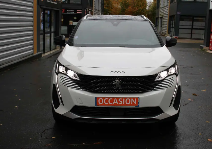 Vue frontale d'un Peugeot 3008 blanc nacré avec calandre noire et phares LED allumés, sur parking humide.