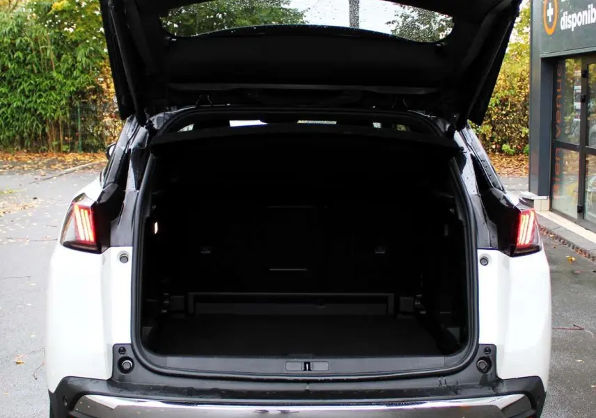 Vue arrière du Peugeot 3008 blanc nacré 2025 avec coffre ouvert et feux arrière allumés sur parking extérieur humide.