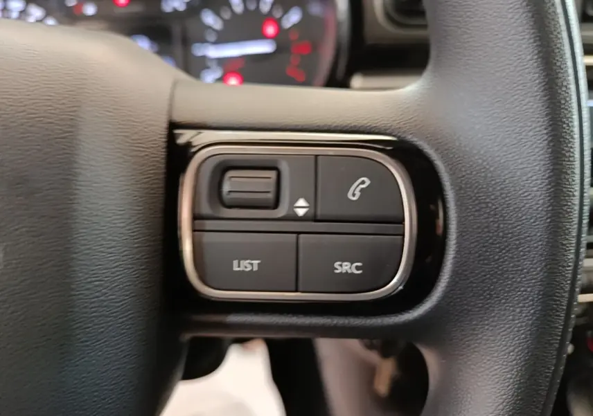 Gros plan sur les commandes multifonctions noires du volant de la Citroën C3 PureTech 110 blanc, avec tableau de bord flou en arrière-plan.