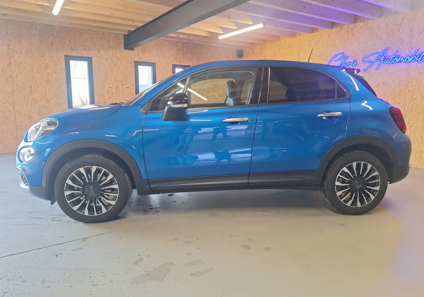Profil côté gauche d'un FIAT 500X bleu métallisé 2024 avec jantes sport et protections noires sur châssis.