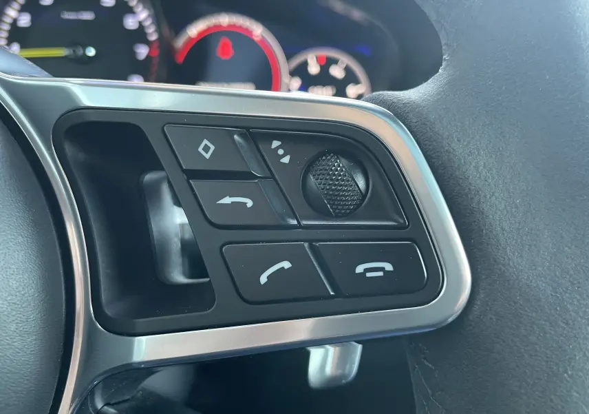 Gros plan sur les commandes au volant à gauche du Porsche Cayenne E-Hybrid gris Kerguelen, avec boutons multifonctions et molette.