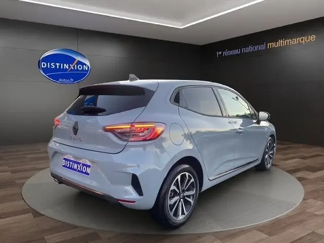 Vue 3/4 arrière droite d'une Renault Clio Techno TCe 90 gris clair, feux LED allumés, sur plateau en showroom.