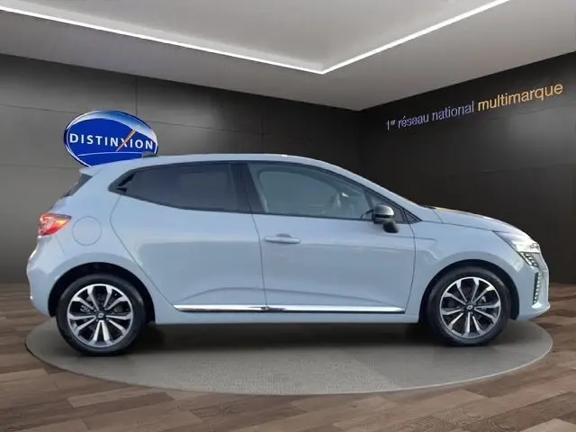 Profil côté gauche d'une Renault Clio Techno TCe 90 gris clair, avec jantes alliage et vitres teintées en arrière-plan showroom.