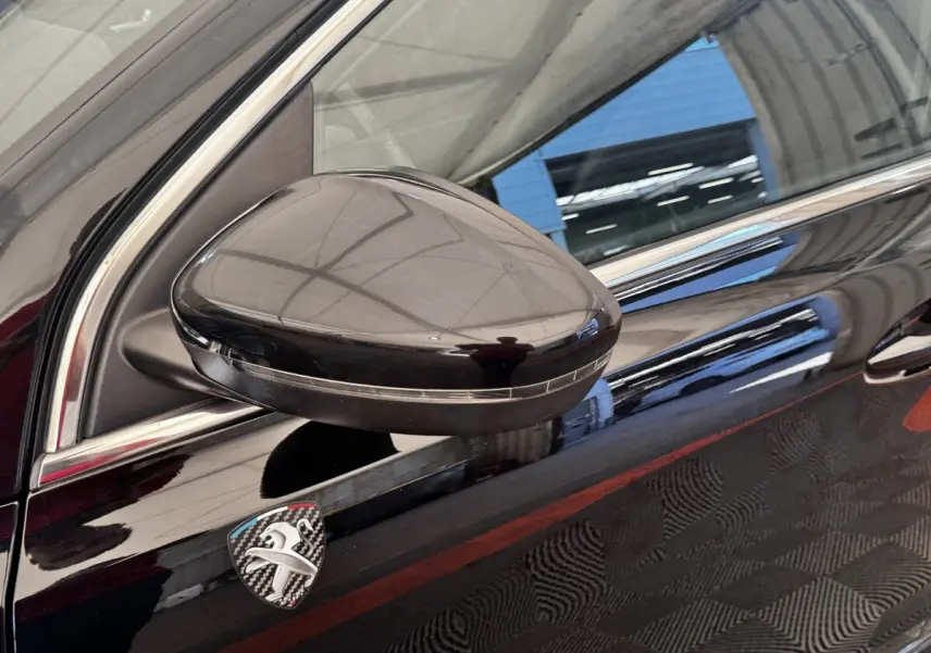 Gros plan sur le rétroviseur côté gauche noir brillant et l'écusson Peugeot en carbone sur la porte noire du Peugeot 308.