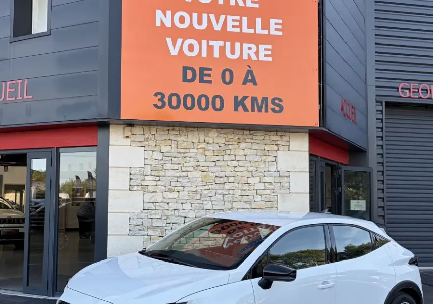 Renault Clio blanche vue en 3/4 avant droit, avec calandre noire et jantes au design moderne, devant un bâtiment commercial.