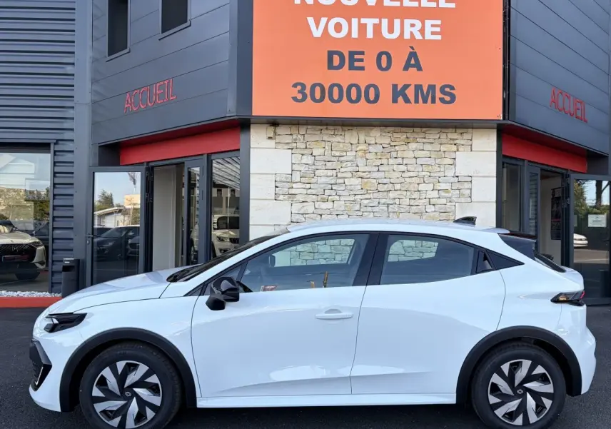 Profil côté gauche d'une Renault Clio blanche 2026 garée devant un bâtiment avec un panneau orange indiquant "votre nouvelle voiture de 0 à 30000 kms".