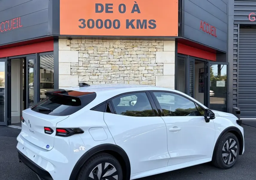 Renault Clio blanche vue 3/4 arrière droit, avec feux arrière LED et jantes au design moderne, devant un bâtiment commercial.