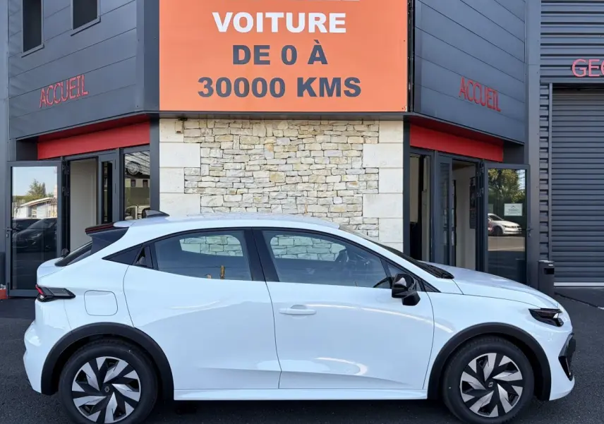 Profil droit d'une Renault Clio blanche 2026 garée devant un bâtiment avec une enseigne orange "Votre nouvelle voiture de 0 à 30000 kms".