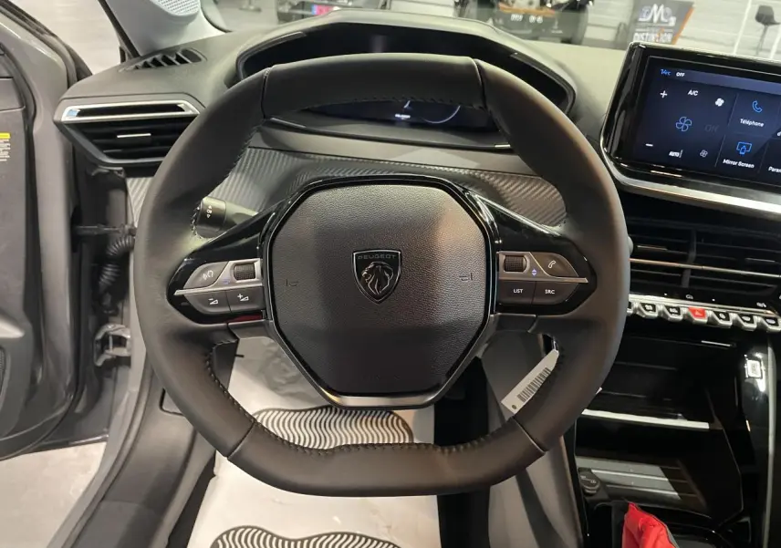 Vue intérieure centrée sur le volant compact cuir de la Peugeot 208 1.2i Hybrid gris Selenium avec tableau de bord numérique.