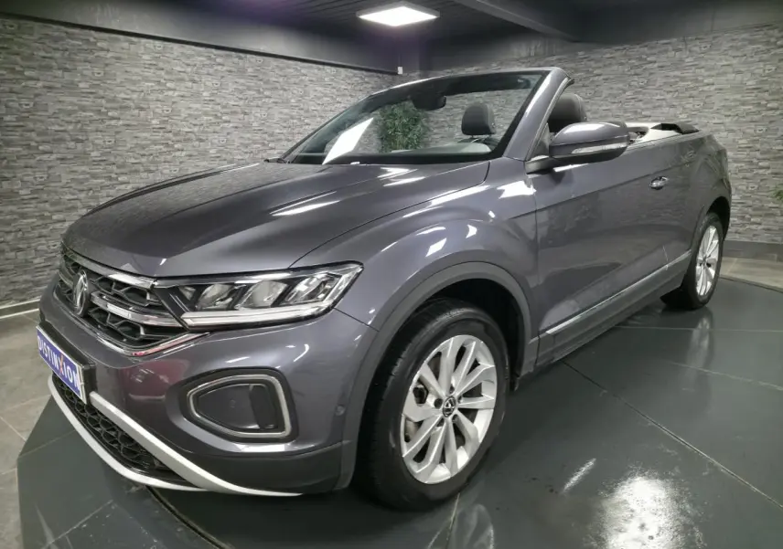 Volkswagen T-Roc cabriolet gris métallisé en 3/4 avant droit, capote ouverte et jantes alliage visibles.