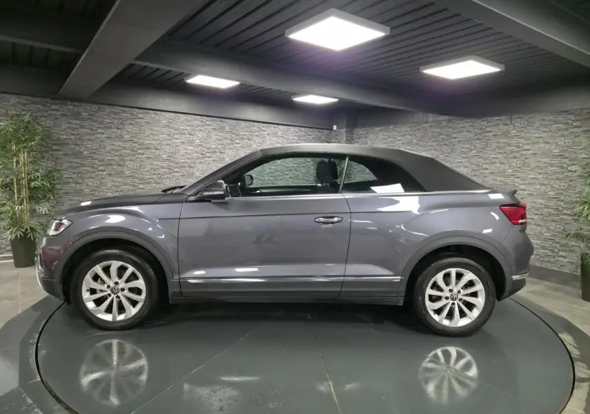 Volkswagen T-Roc Cabriolet gris métallisé vue de profil côté gauche avec capote noire fermée en intérieur showroom.