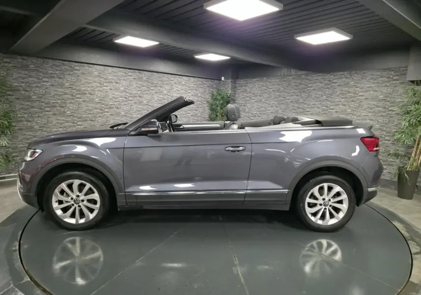 Vue de profil côté gauche d'un Volkswagen T-Roc Cabriolet gris métallisé avec capote ouverte en intérieur showroom.