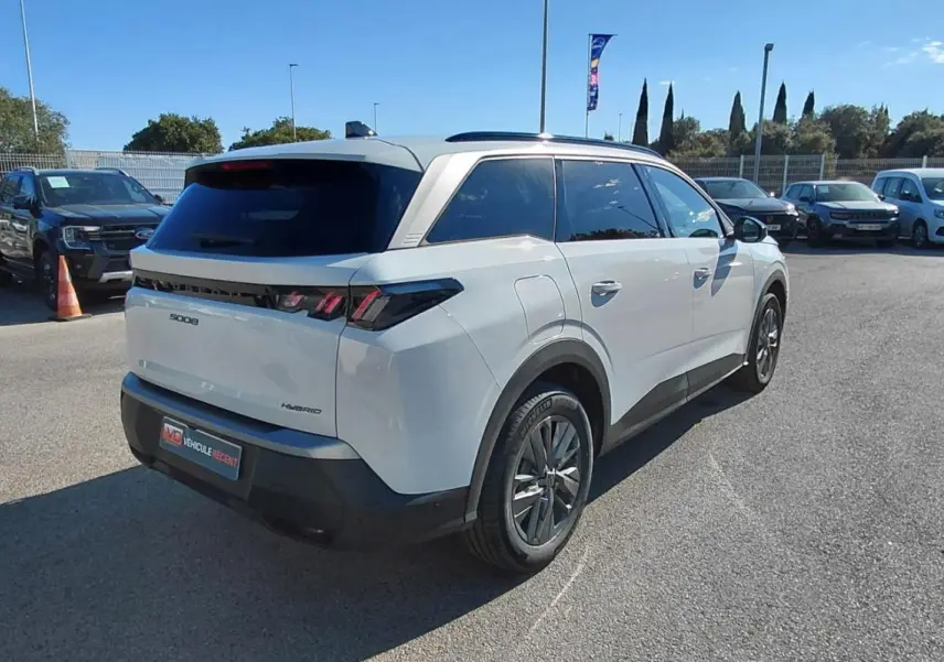 Peugeot 5008 Hybrid blanc vu en 3/4 arrière droit sur parking, avec feux arrière à griffes distinctifs et toit panoramique.
