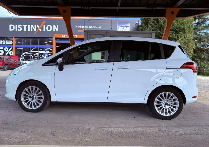 Profil côté gauche d'une Ford B-MAX blanche de 2014 avec jantes alliage et vitres teintées, stationnée sous abri.