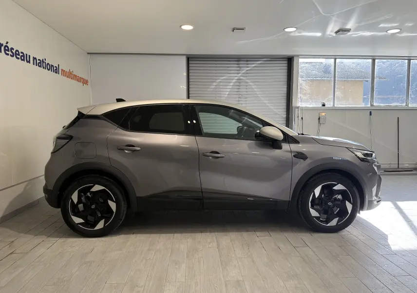 Profil droit du Renault Captur 2025 gris Cassiopée métal avec toit gris, roues noires à jantes stylisées, en intérieur showroom.