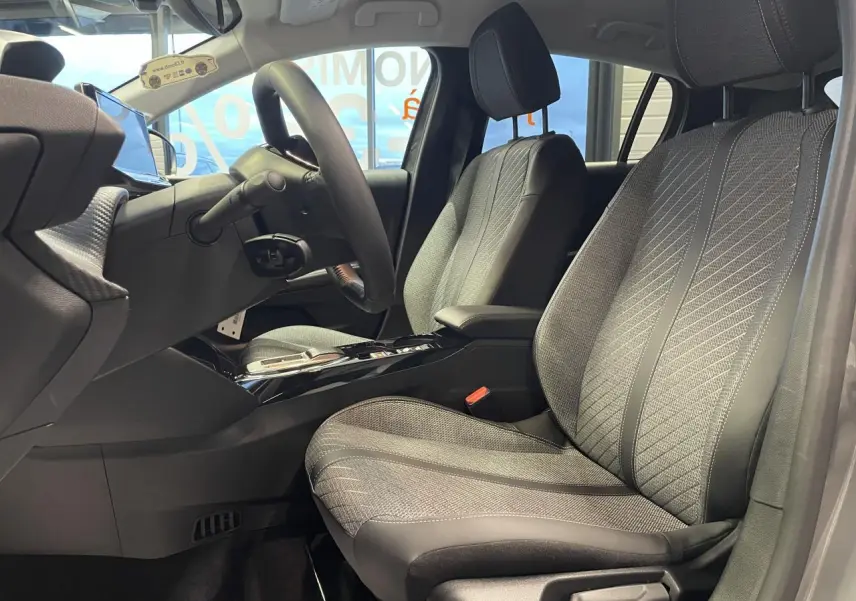 Vue intérieure côté gauche des sièges avant gris clair et noir de la Peugeot 208 1.2i Hybrid 2025 avec accoudoir central.