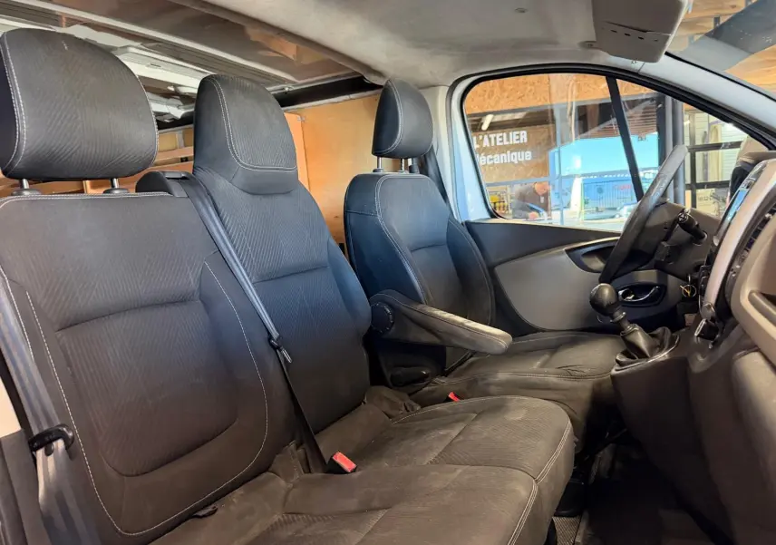 Vue intérieure côté conducteur du Renault Trafic Fourgon blanc, montrant les sièges noirs et le levier de vitesses manuel.