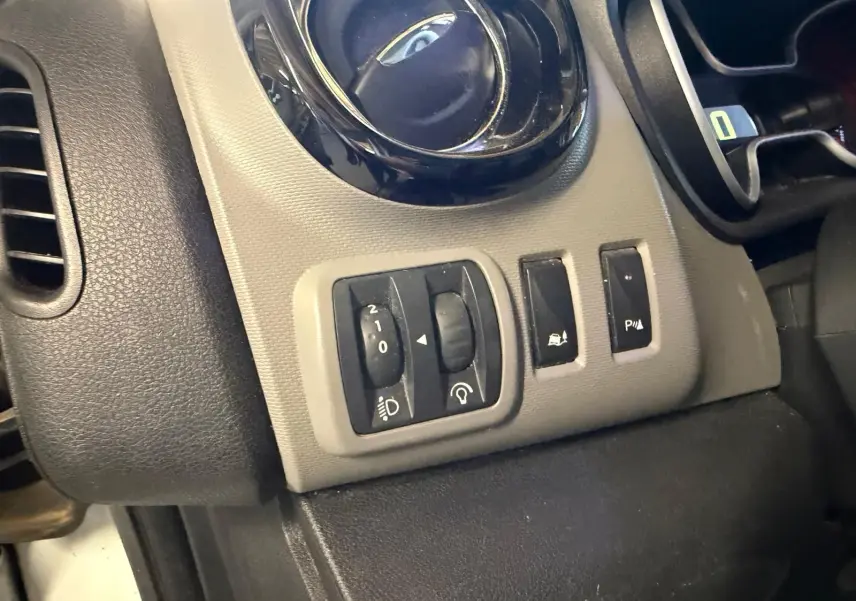 Gros plan sur les commandes de réglage des phares et aides à la conduite du tableau de bord gris et noir du Renault Trafic blanc.