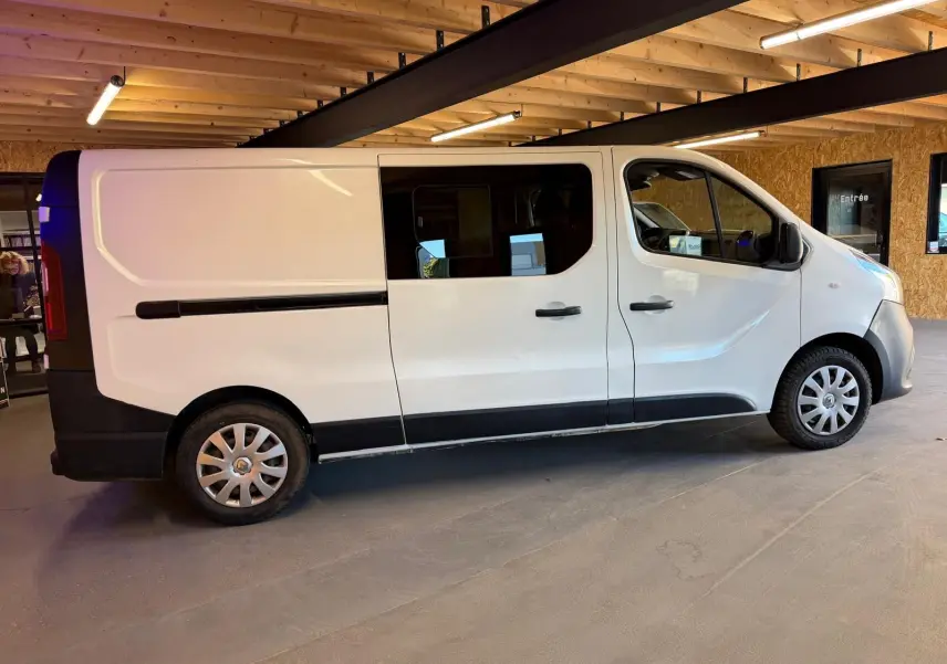 Vue de profil côté droit d'un Renault Trafic Fourgon blanc avec fenêtres latérales et jantes argentées, stationné en intérieur.