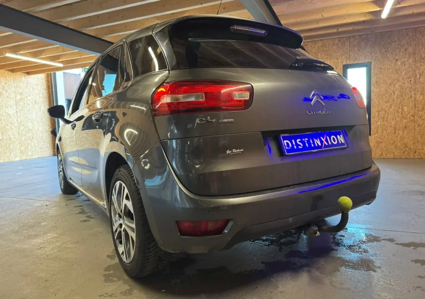 Vue 3/4 arrière gauche d'une Citroën C4 Picasso gris métallisé avec attelage et jantes alliage en intérieur garage.