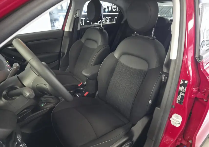 Vue intérieure côté conducteur de la Fiat 500X 2024 rouge, sièges tissu noir avec monogramme FIAT et accoudoir central.