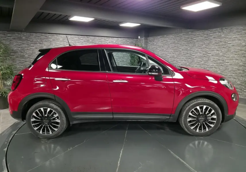 Profil droit d'une Fiat 500X rouge Passion avec jantes alliage 17'' et rétroviseurs noirs dans un showroom.