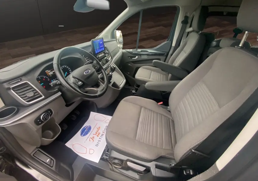 Vue intérieure côté conducteur du Ford Tourneo Custom gris magnetic, montrant le tableau de bord et les sièges avant en tissu gris.