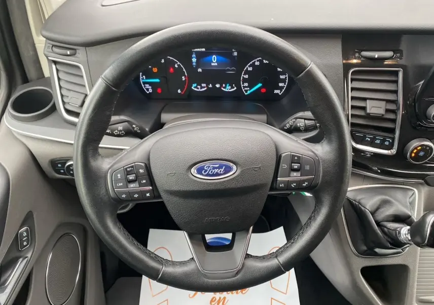 Vue rapprochée du volant noir et du tableau de bord numérique du Ford Tourneo Custom gris Magnetic, avec levier de vitesses manuel.