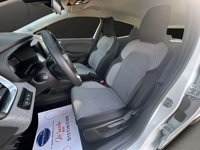 Vue intérieure côté conducteur d'une Renault Clio blanche 2025, sièges tissu gris et volant multifonctions cuir synthétique.