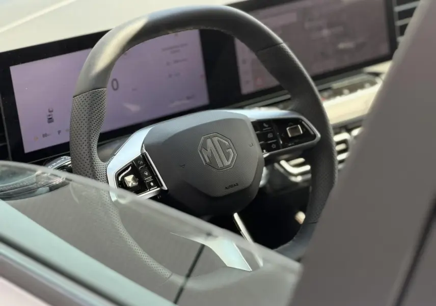 Gros plan sur le volant noir perforé avec logo MG, tableau de bord numérique et écran tactile en arrière-plan.