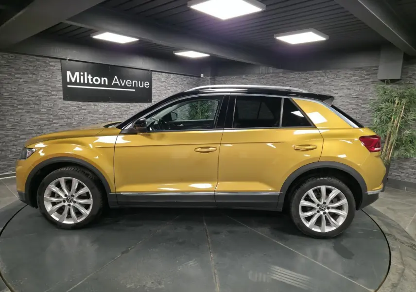 Vue latérale droite d'un Volkswagen T-Roc jaune Curcuma avec toit noir et jantes alliage 18 pouces dans un showroom.