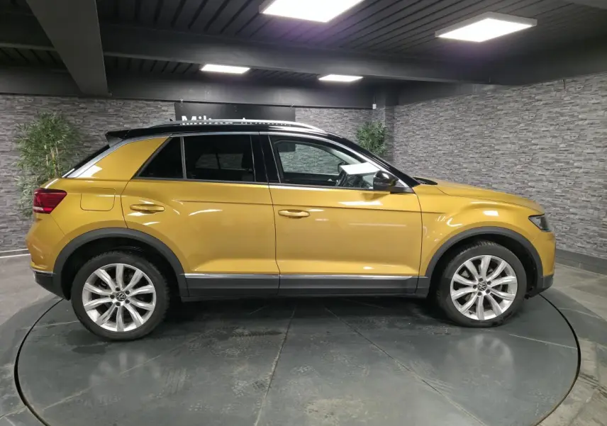 Vue de profil côté gauche d'un Volkswagen T-Roc jaune Curcuma avec toit noir et jantes alliage 18 pouces en intérieur showroom.