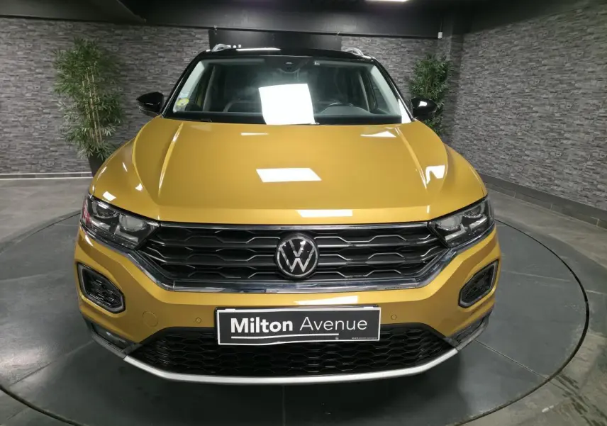 Vue avant d'un Volkswagen T-Roc jaune Curcuma avec calandre noire et détails chromés en intérieur showroom.