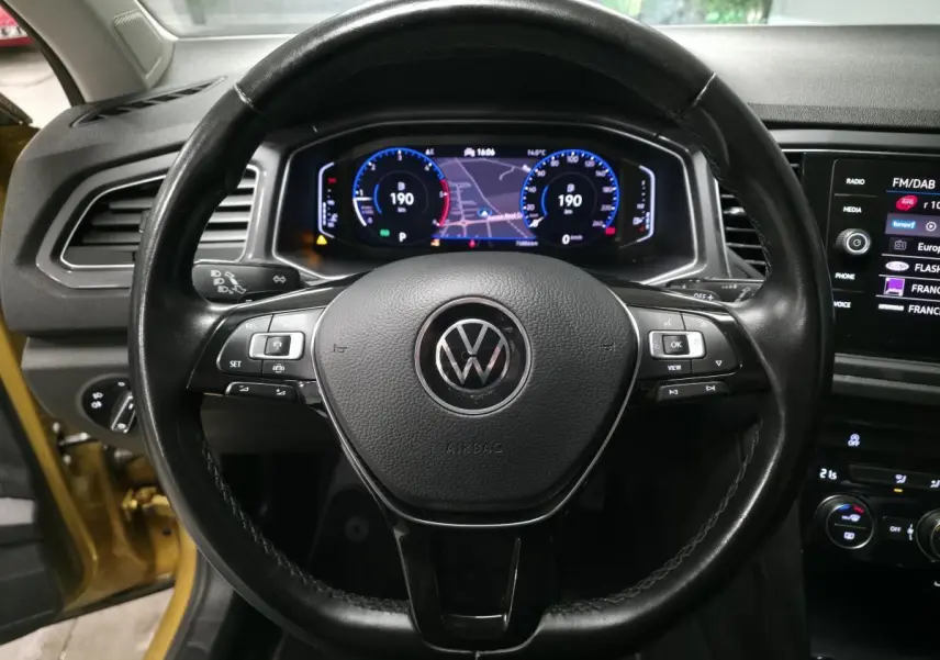 Vue centrée sur le volant en cuir noir d’un Volkswagen T-Roc jaune, avec tableau de bord numérique et écran tactile visible.