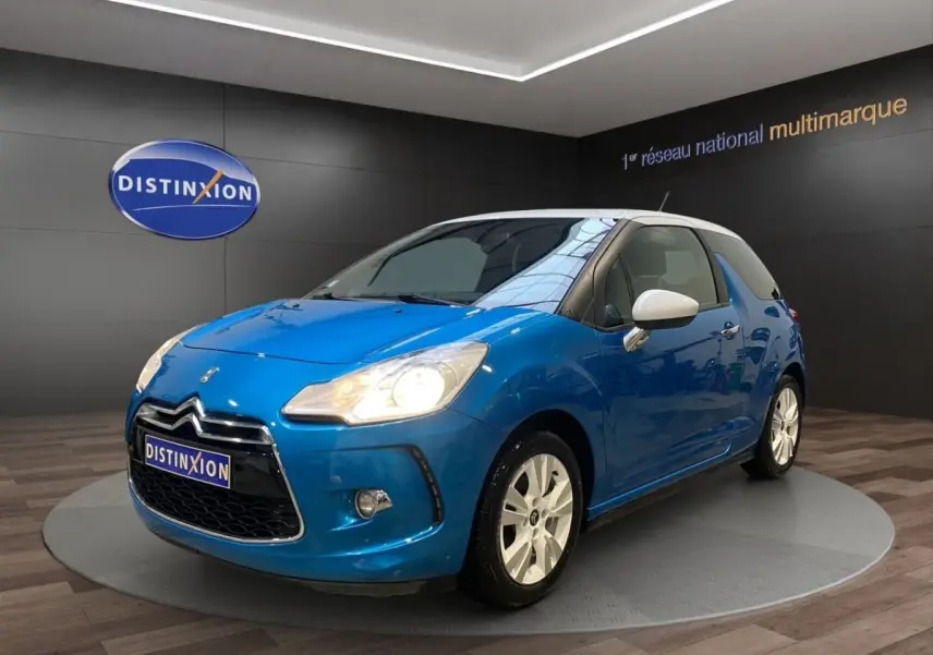 Citroën DS3 e-HDi 90 bleu vue 3/4 avant droit avec toit et rétroviseurs blancs, phares allumés en showroom.