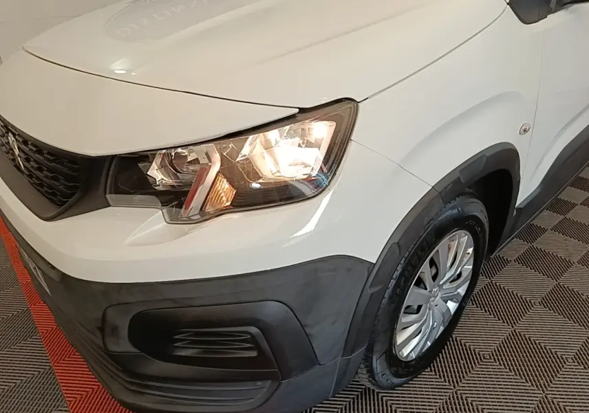 Vue 3/4 avant droit du Peugeot Rifter blanc avec phares allumés et pare-chocs noir mat contrastant.