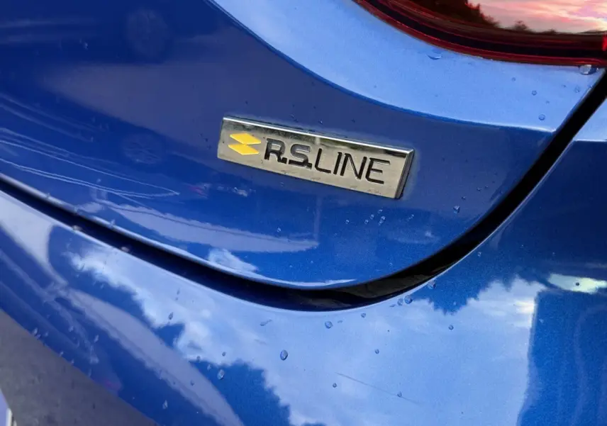 Gros plan sur l'arrière bleu métallisé d'une Renault Clio RS Line avec le badge RS Line visible sous la pluie.