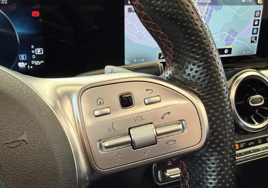Gros plan sur les commandes argentées du volant en cuir noir perforé du Mercedes GLA 250 e AMG Line 2023, avec écran tactile en arrière-plan.