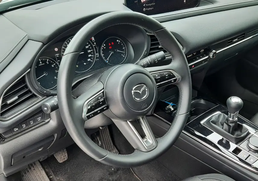 Vue intérieure côté conducteur de la Mazda CX-30 2024, volant cuir, écran central, boîte manuelle et tableau de bord noir.