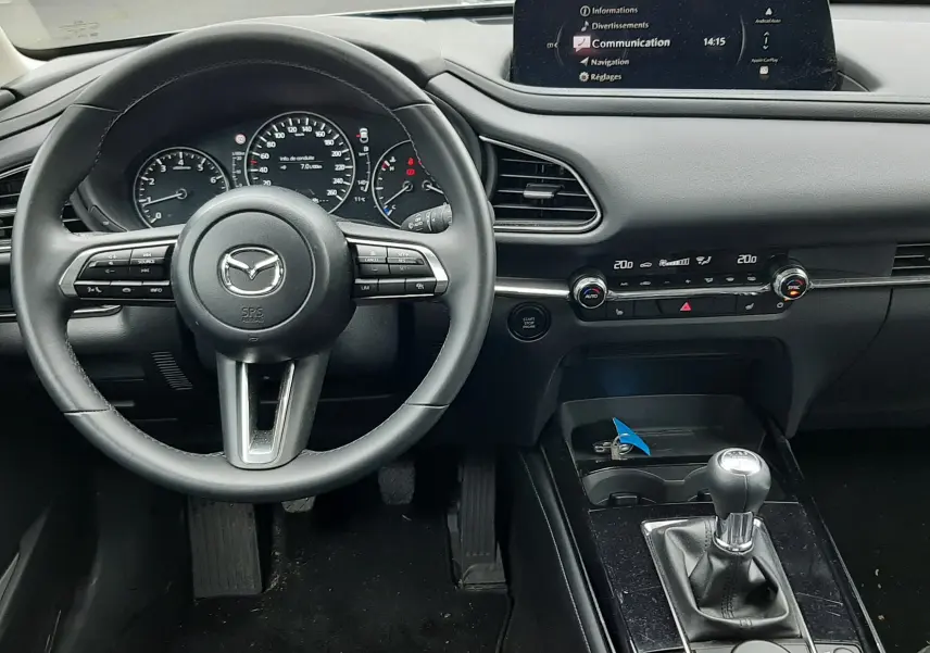 Vue intérieure du poste de conduite du Mazda CX-30 2024, volant cuir, tableau de bord noir et levier de vitesse manuel.
